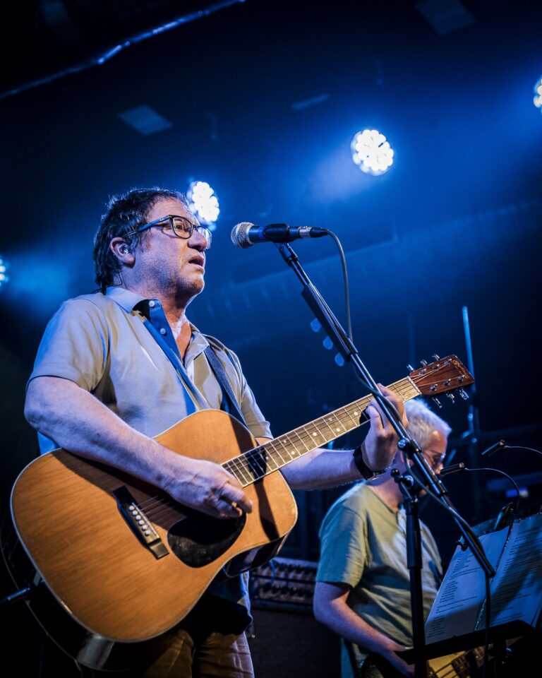 Simon Fowler cantando con la guitarra acústica bajo luz azul y el bajista detrás en la Sala Custom de Sevilla