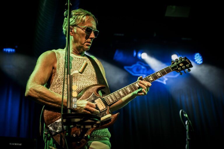 Steve Cradock tocando la guitarra con chaleco de punto y gafas de sol bajo luz azul en la Sala Custom de Sevilla